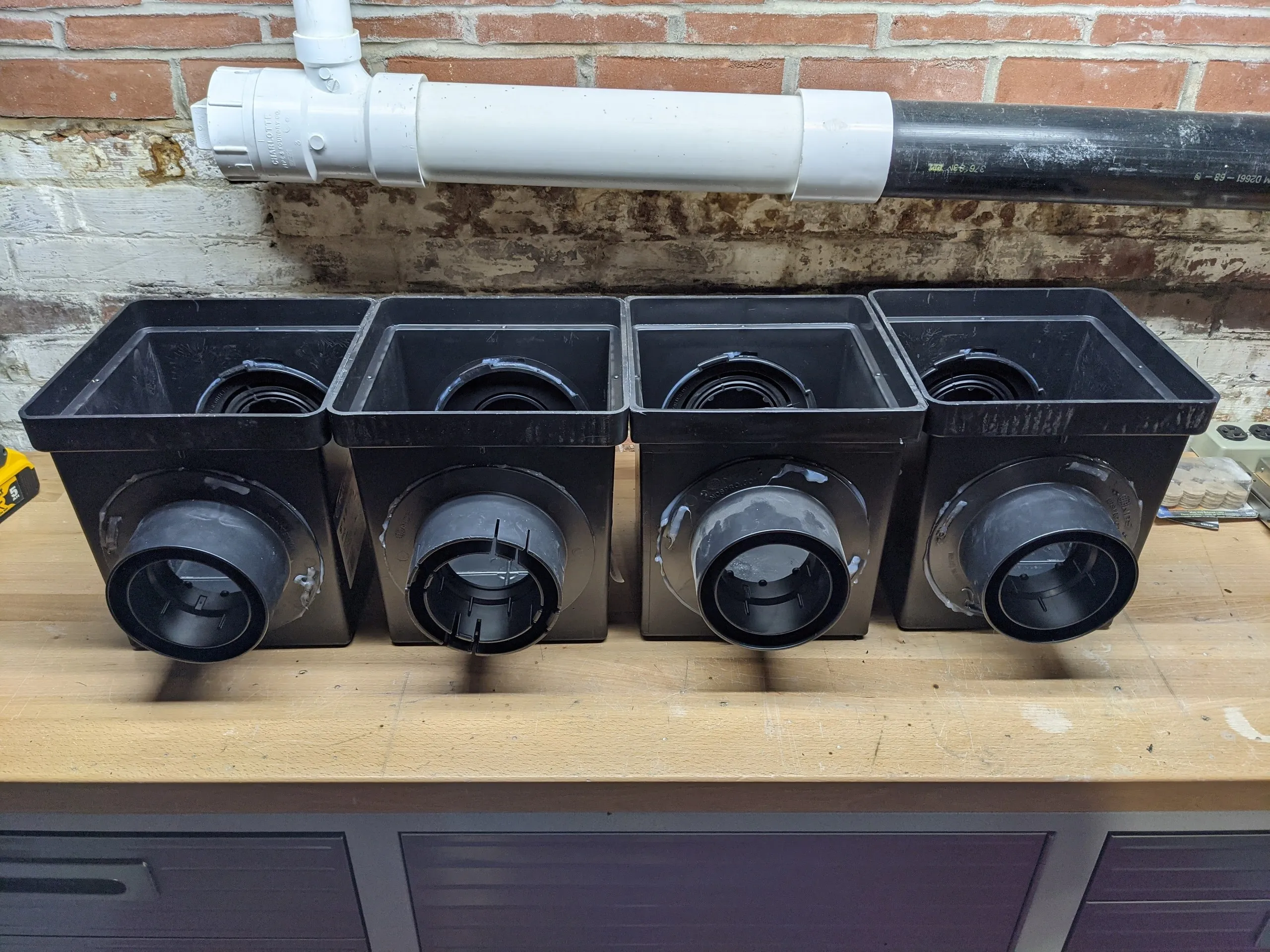 Four catch basins on a workbench, each with a corrugated pipe outlet solvent-welded and sealed with silicone