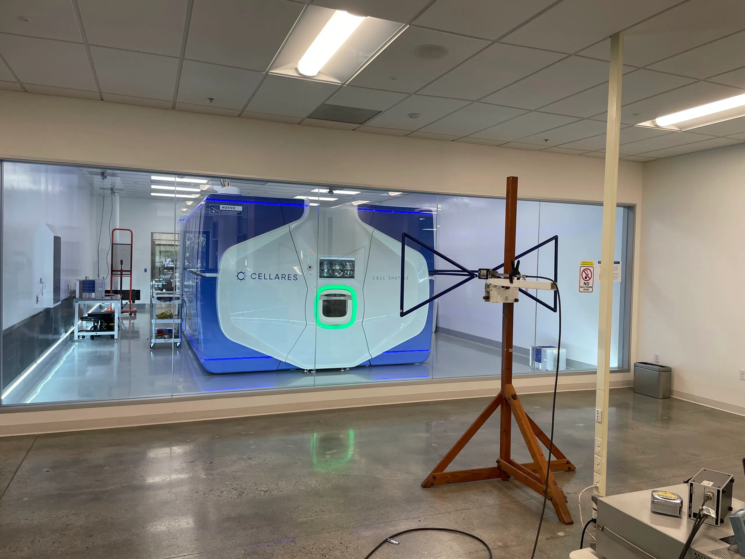 The Cell Shuttle viewed through a cleanroom window, with an antenna on a wooden stand in the foreground for EMC testing