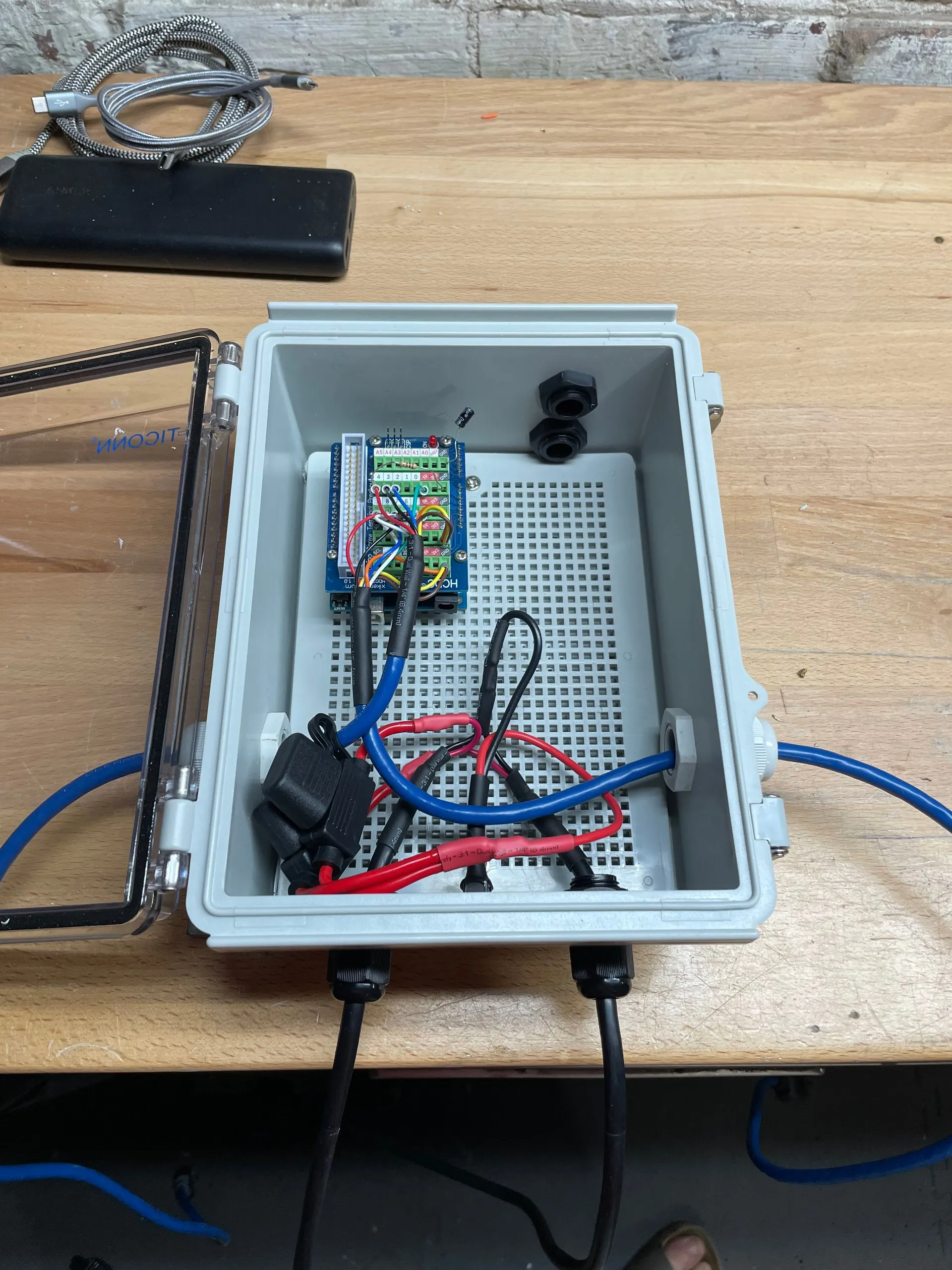 A grey sealed enclosure open on a bench, containing a small breakout board on a mounting plate with red and blue battery leads and an inline fuse