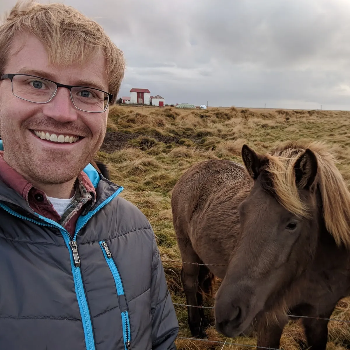 Darren Brown outdoors in a grey and blue jacket, smiling beside an Icelandic horse at a wire fence, with a red-roofed farm building behind