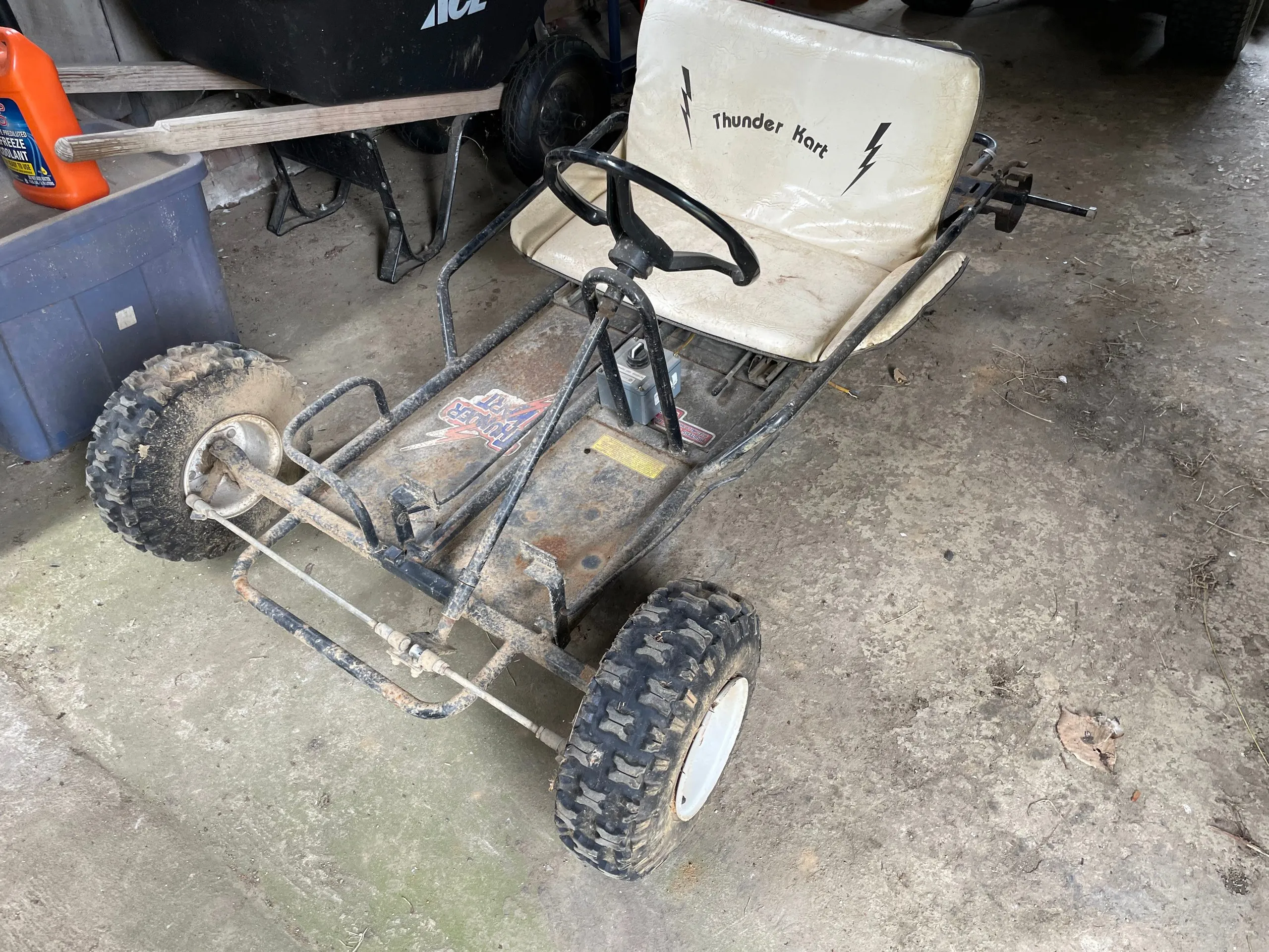 The Thunder Kart in its original state, a rusty steel frame with a white vinyl seat, steering wheel and four knobby tyres, parked on a garage floor