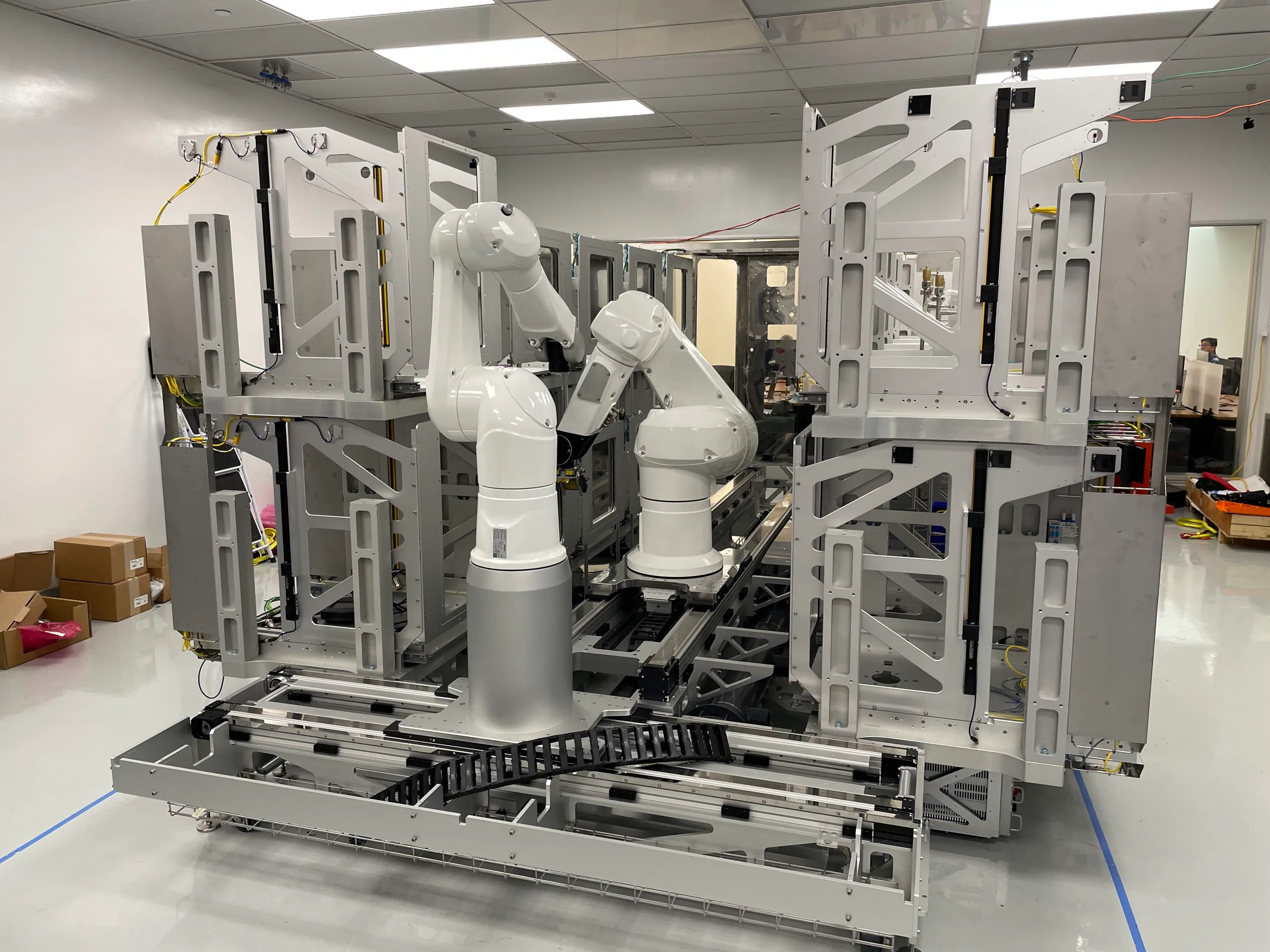 Two articulated robot arms mounted on a mobile chassis, positioned inside a large stainless steel and aluminum frame with sliding doors on either side