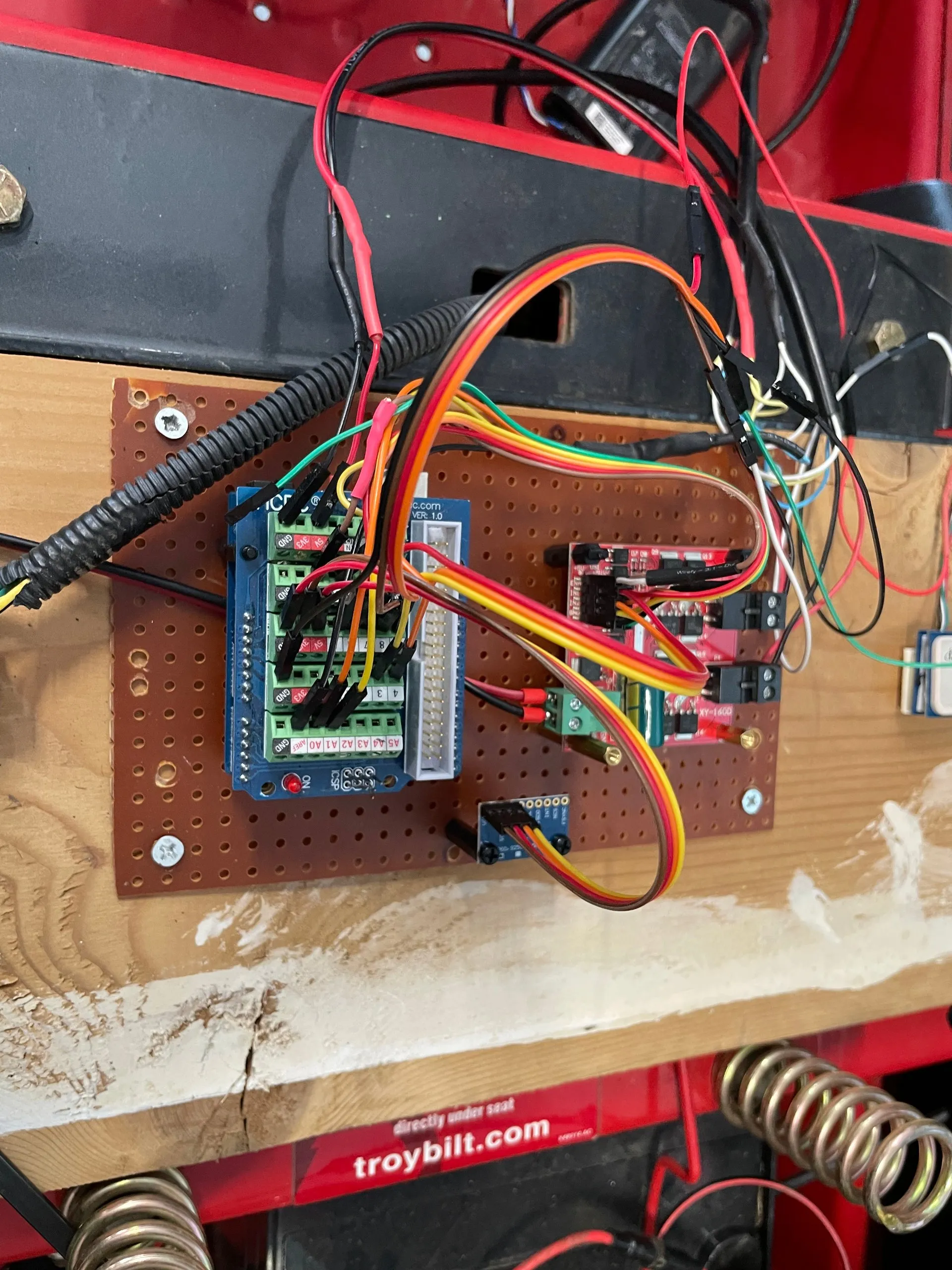 A perfboard mounted under the mower’s seat rail carrying a screw-terminal Arduino shield, a dual H-bridge motor driver and ribbon-wired connections to the actuators and sensors