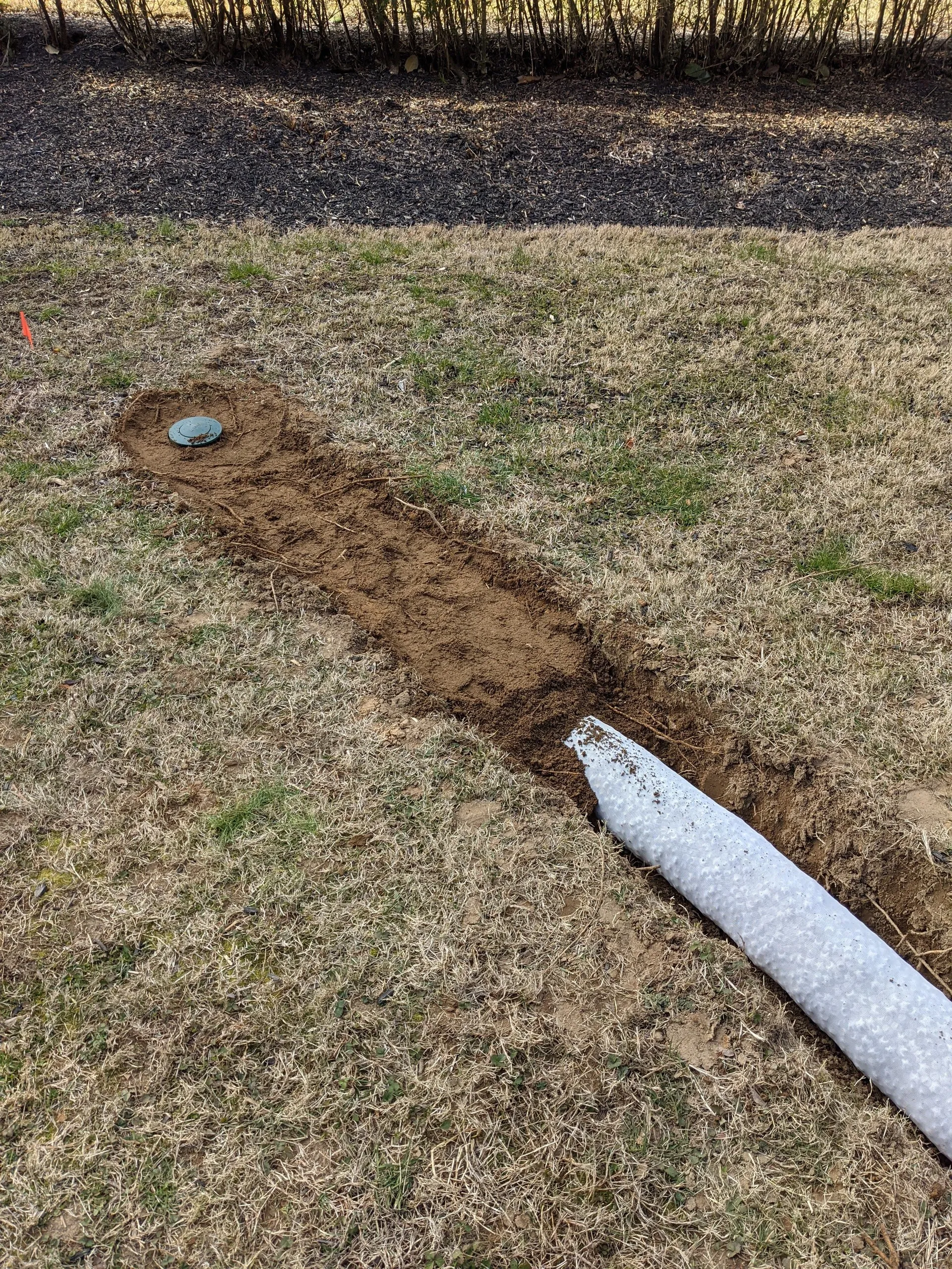 Open trench in the lawn leading to a round pop-up emitter cap, with the sock-wrapped pipe visible at the near end