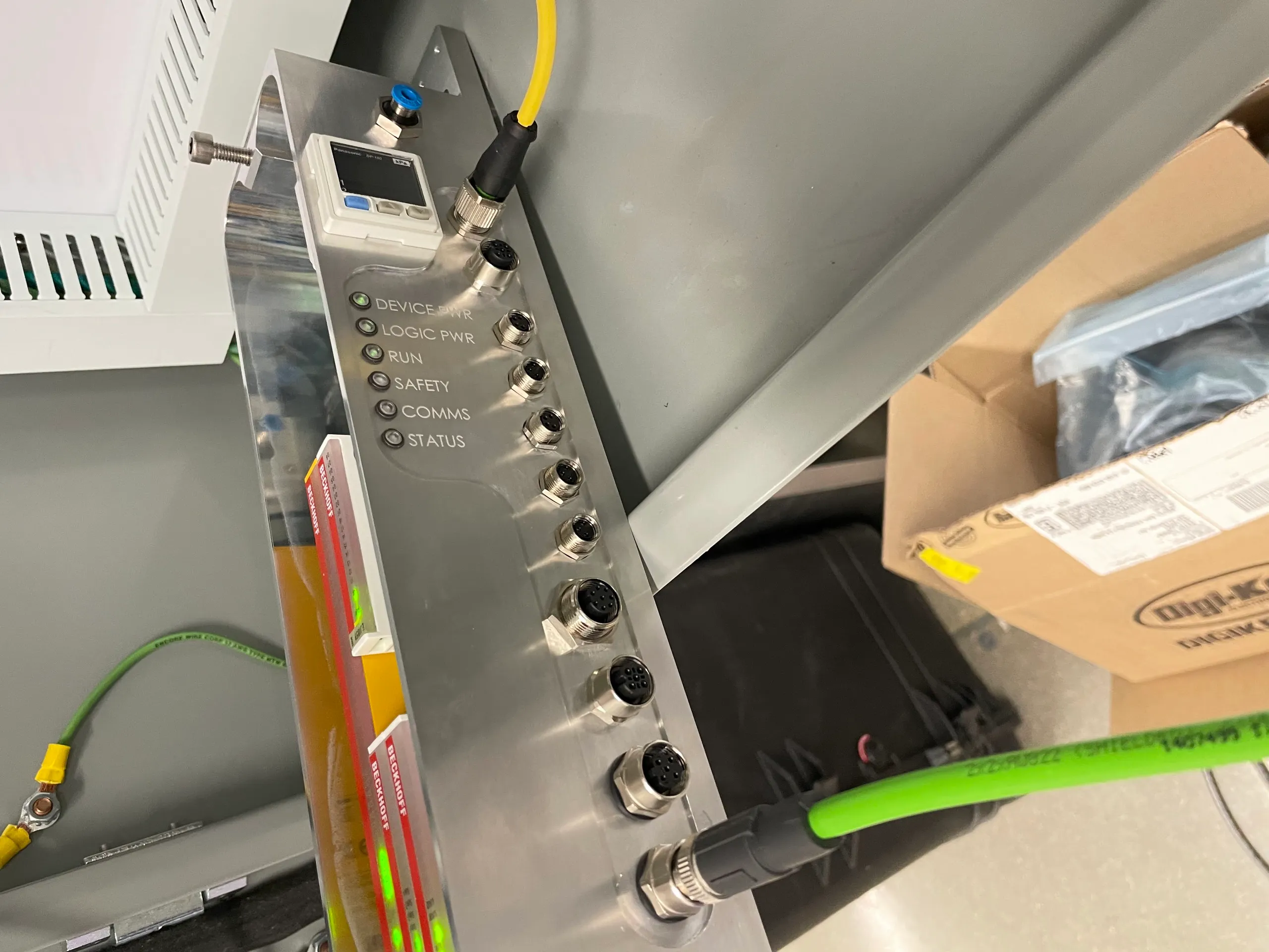 The same interface board mounted in a stainless steel panel, with M12 connectors, a small digital pressure gauge, and status LEDs labeled Device Pwr, Logic Pwr, Run, Safety, Comms, and Status
