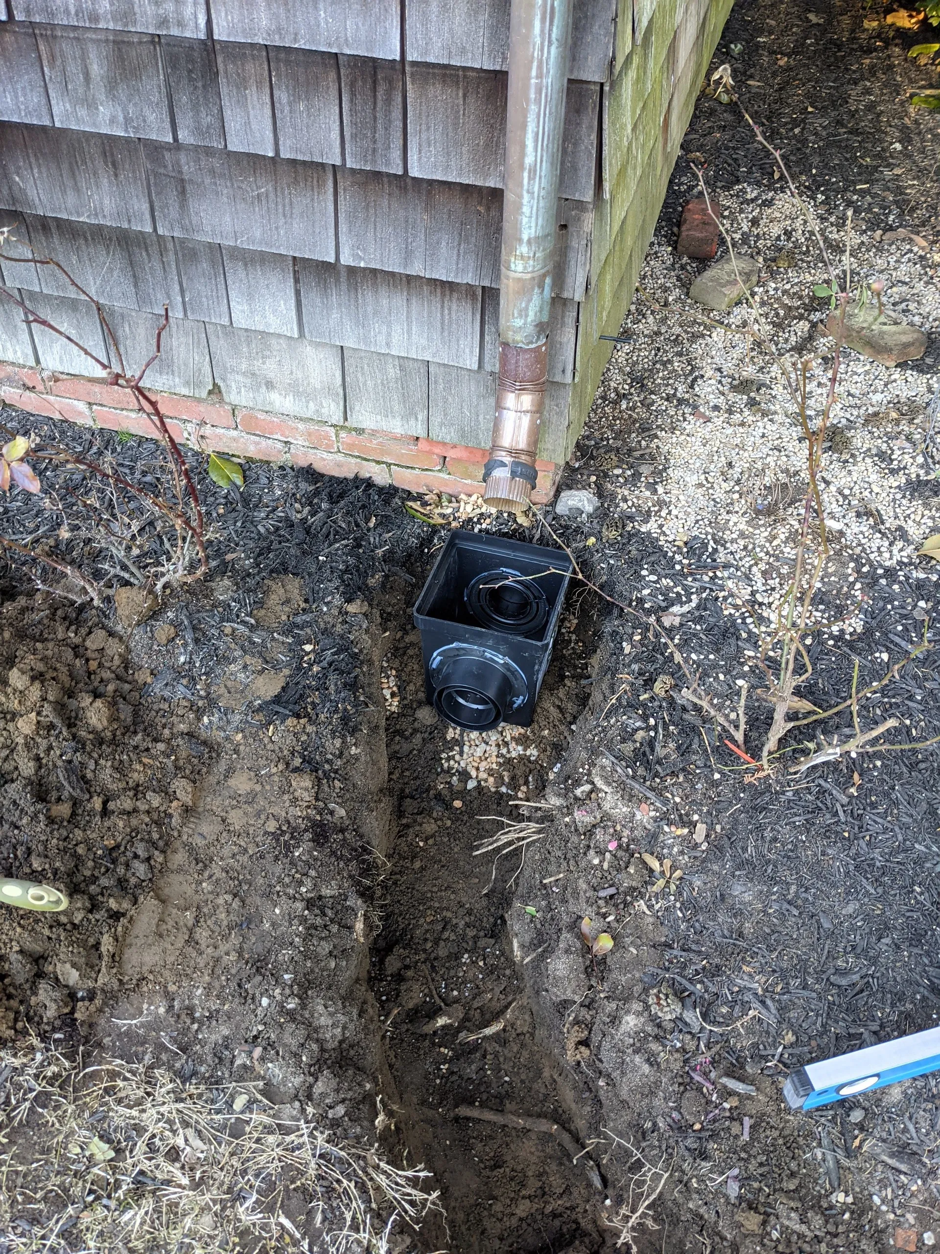 Catch basin set in the ground under a copper downspout, at the corner of the house, with the connecting trench dug alongside it