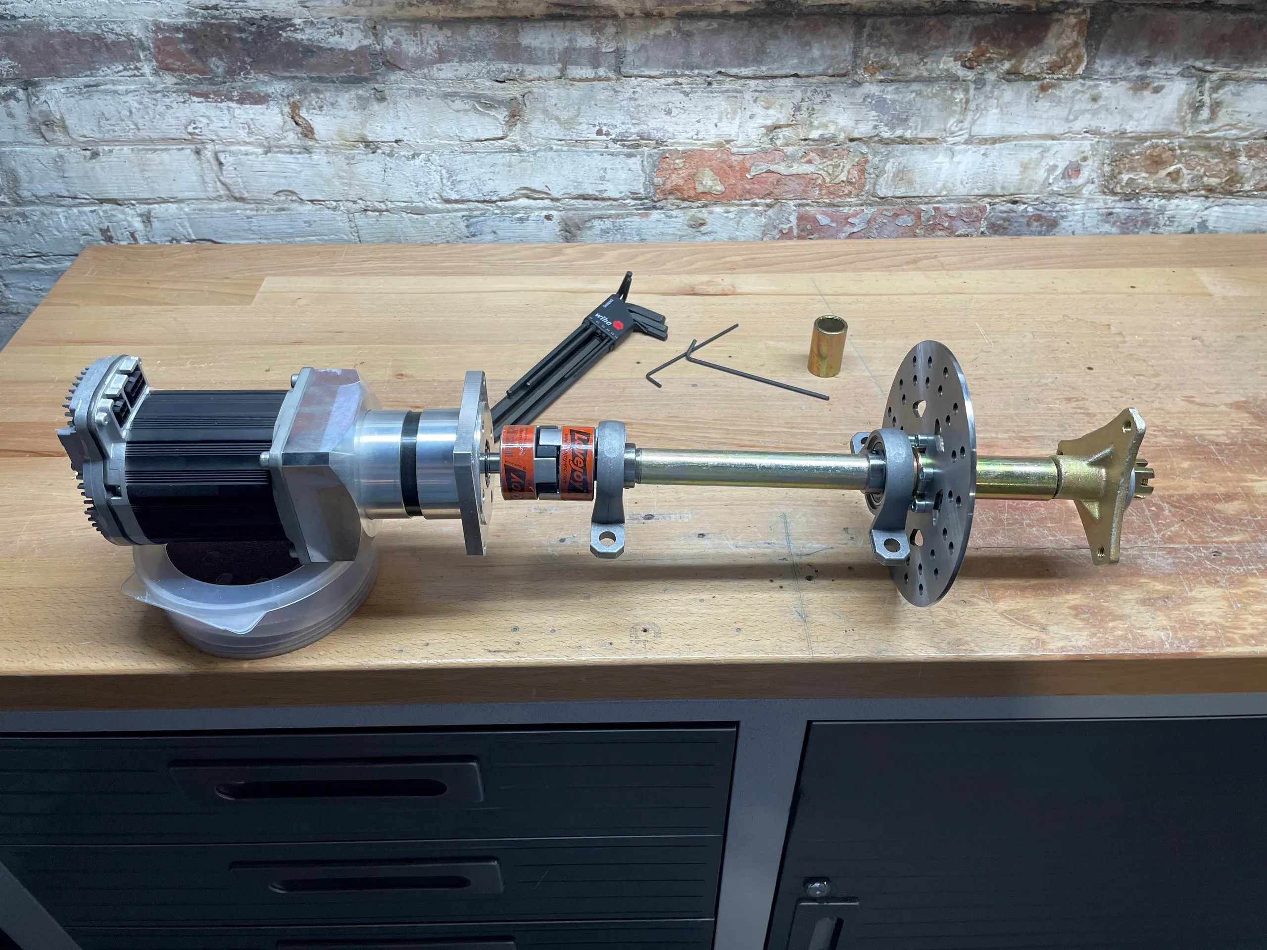 A complete drive assembly laid out on a workbench: motor, gearbox, coupling, shaft in two pillow blocks, brake disc and wheel hub