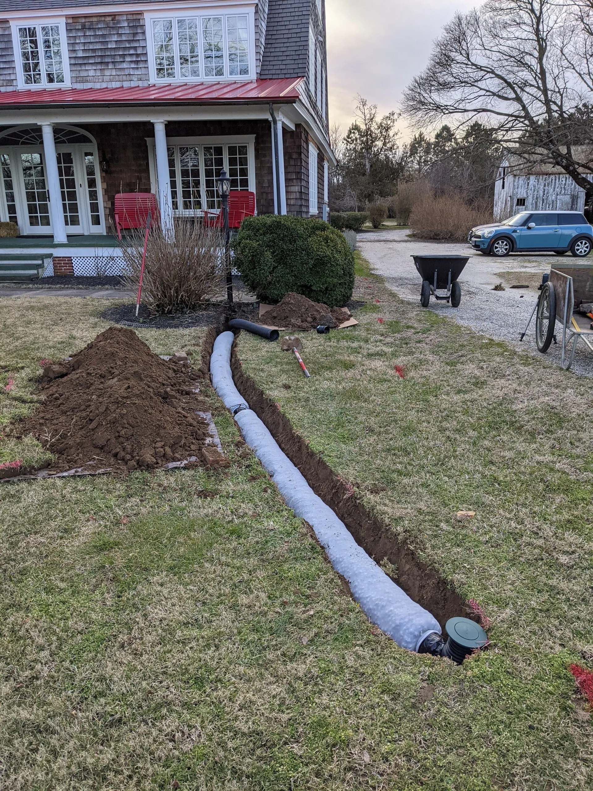 Long curving trench across the front lawn with sock-wrapped pipe leading toward a pop-up emitter, house and driveway in the background