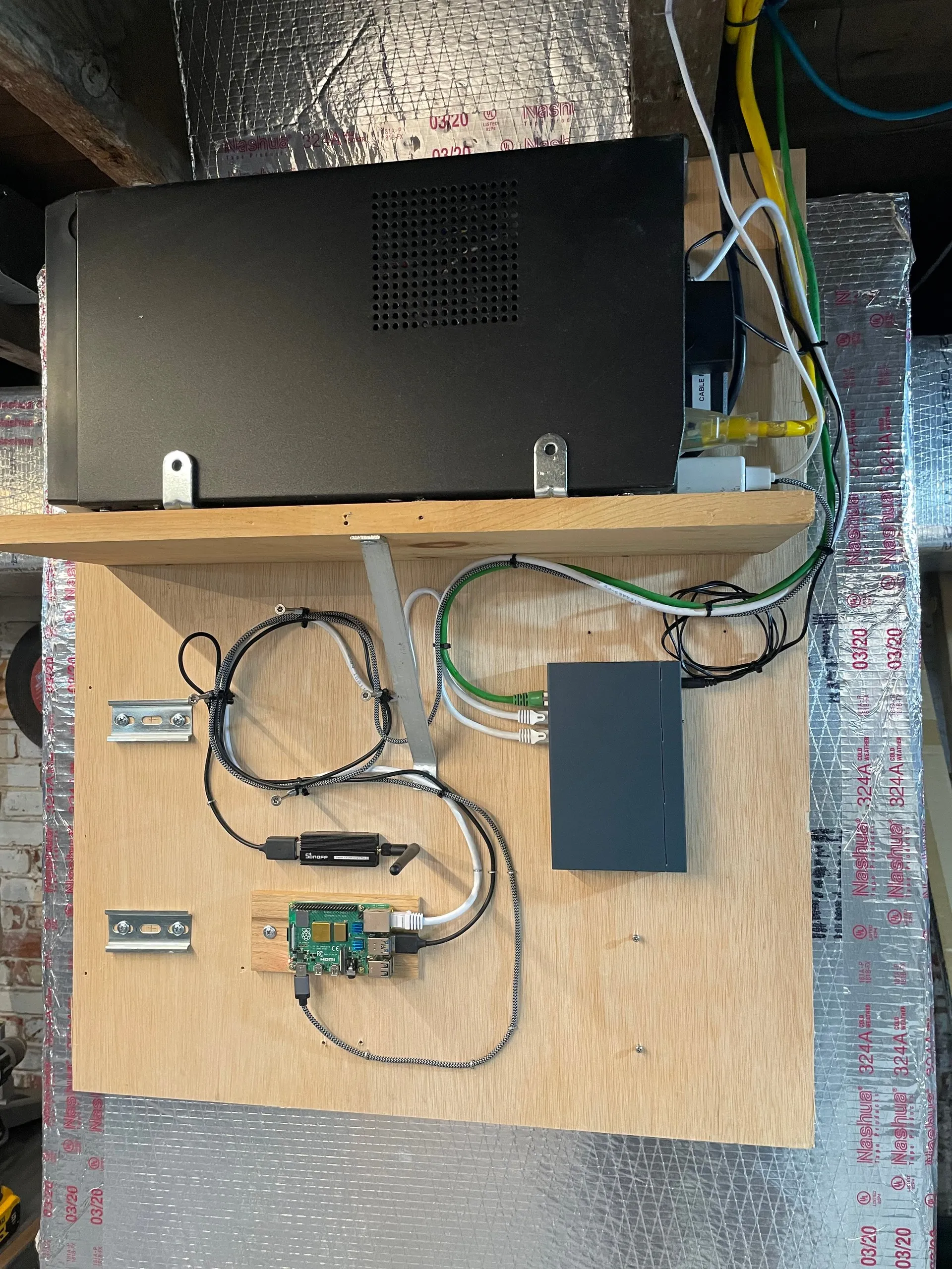 A Raspberry Pi 4 and a Sonoff Zigbee USB stick mounted on a plywood panel in the crawlspace, with a network switch beside them and a rack-mount APC UPS on the shelf above
