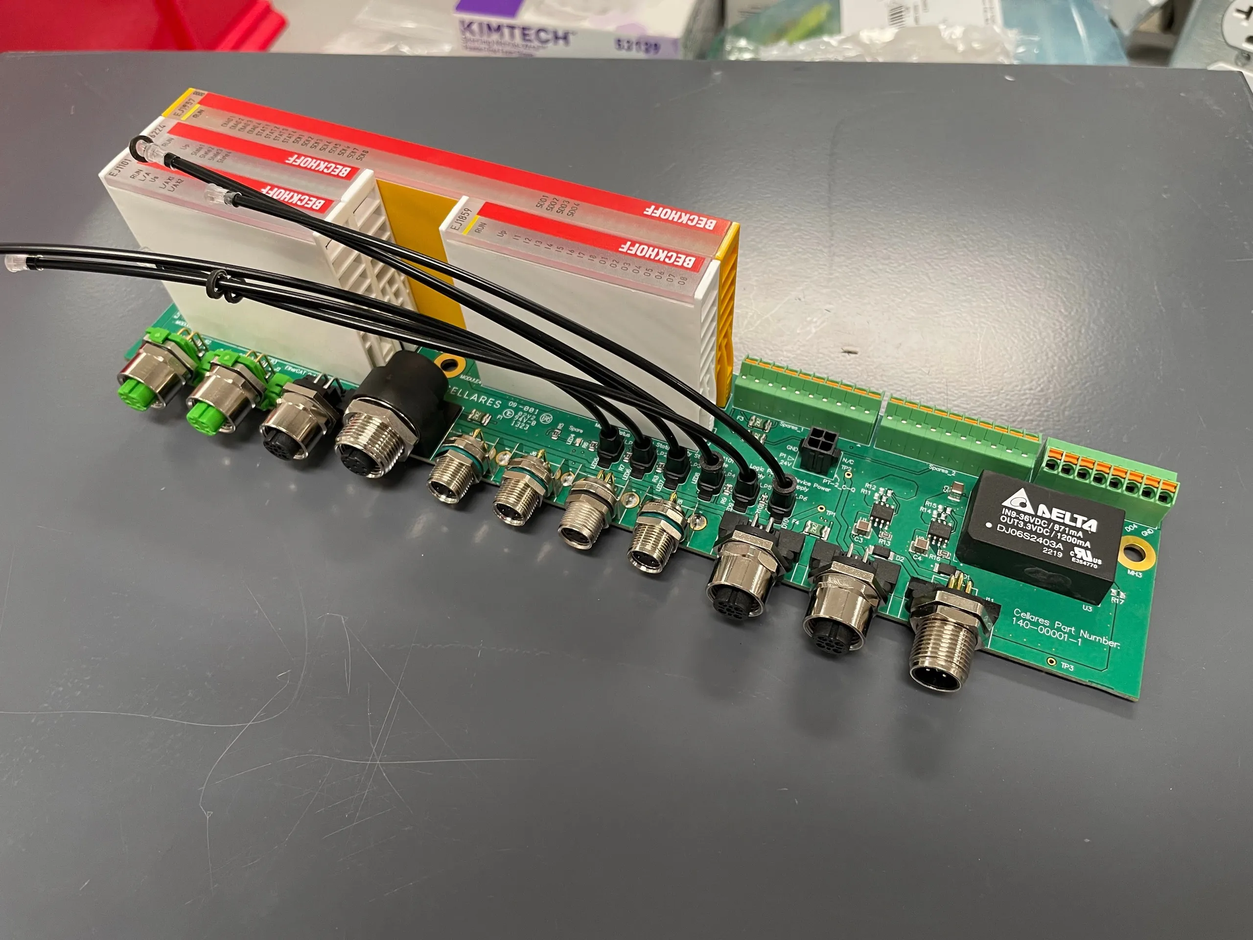 A green custom-designed circuit board with M12 connectors along one edge, connected by short cables to two white Beckhoff EtherCAT terminal blocks standing on end