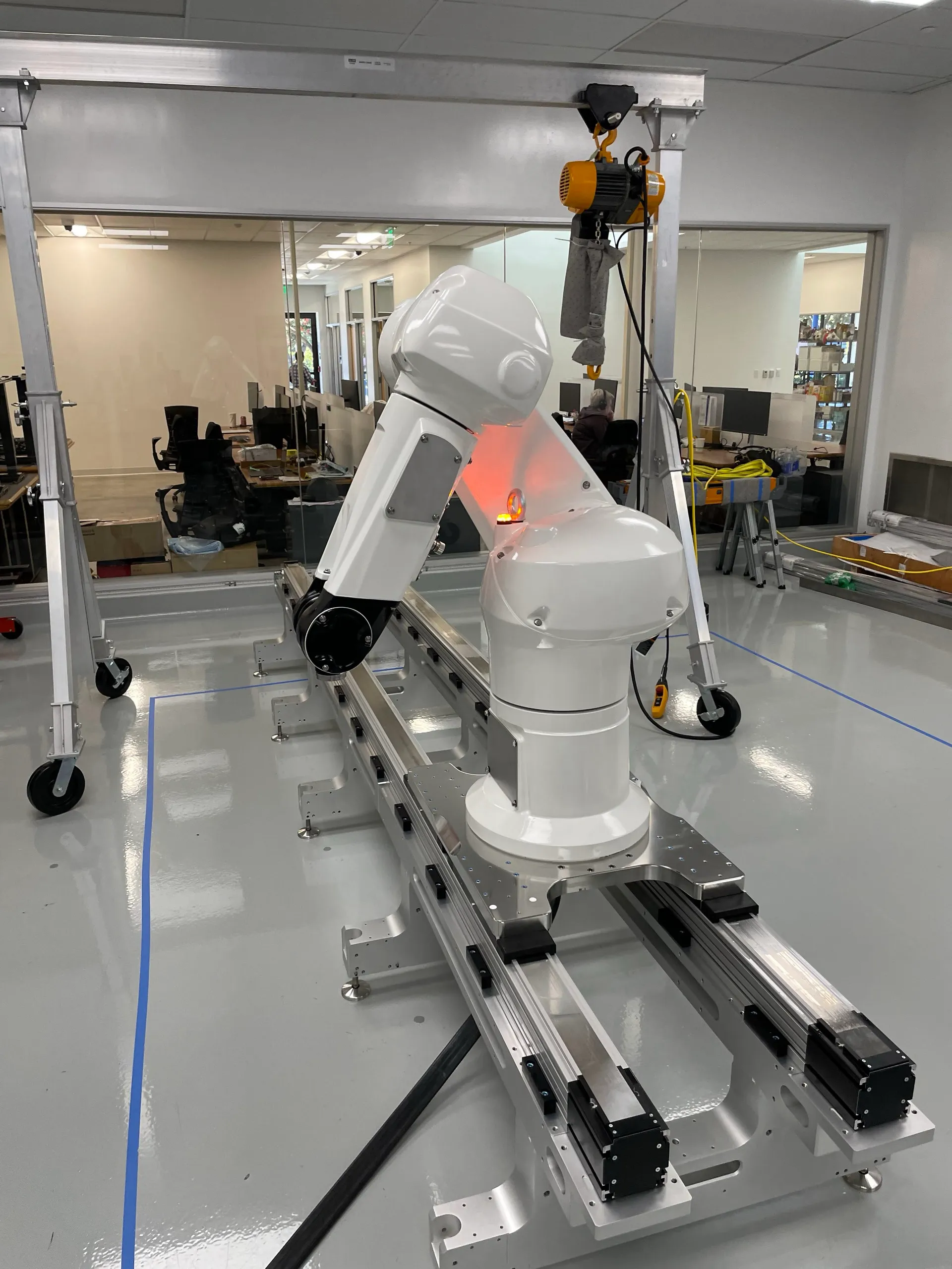 A single white robot arm mounted on a linear rail inside a lab, with a chain hoist positioned above it