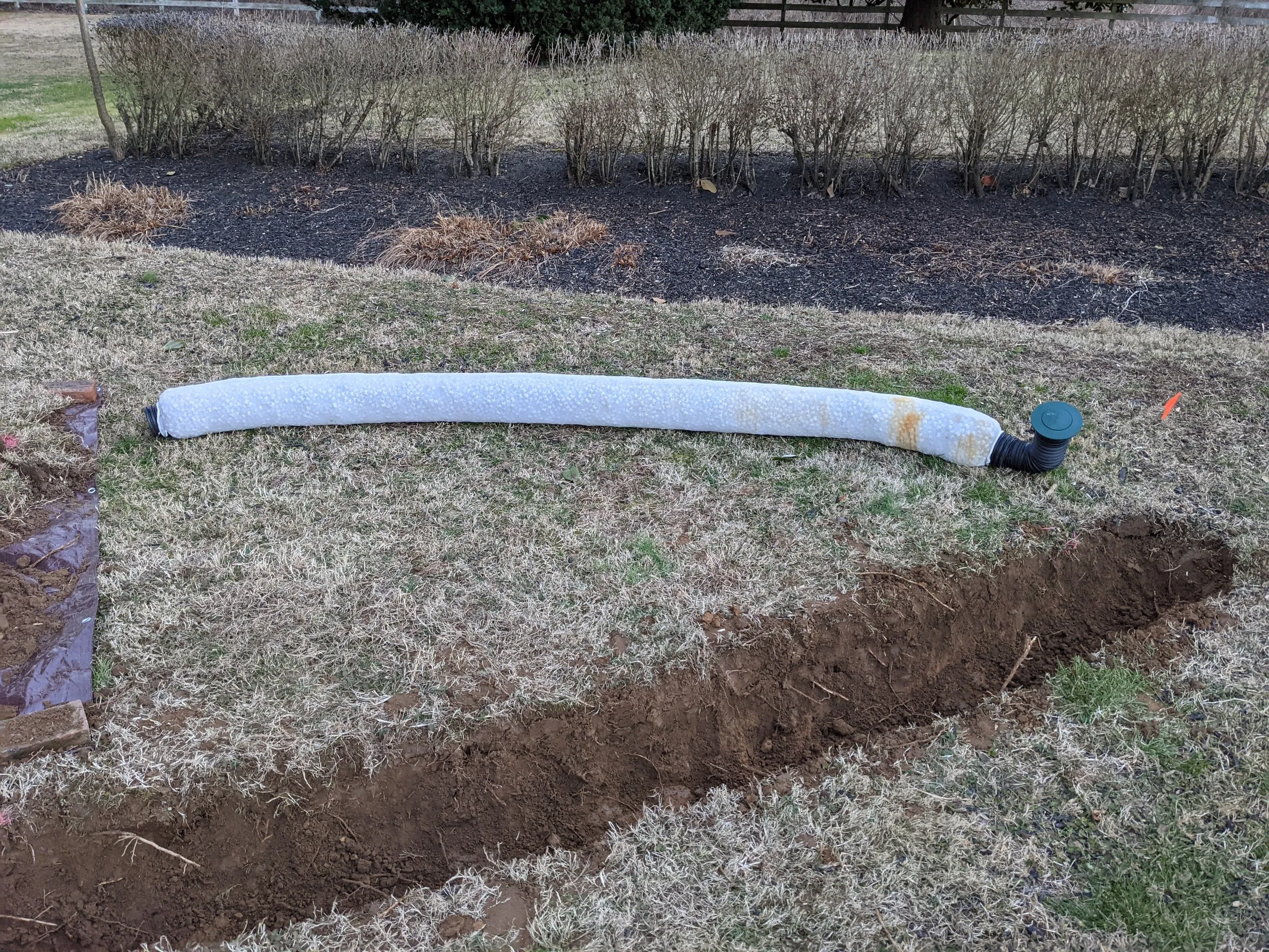 Corrugated pipe wrapped in a fabric sock lying in the grass, running from the trench to a green pop-up emitter