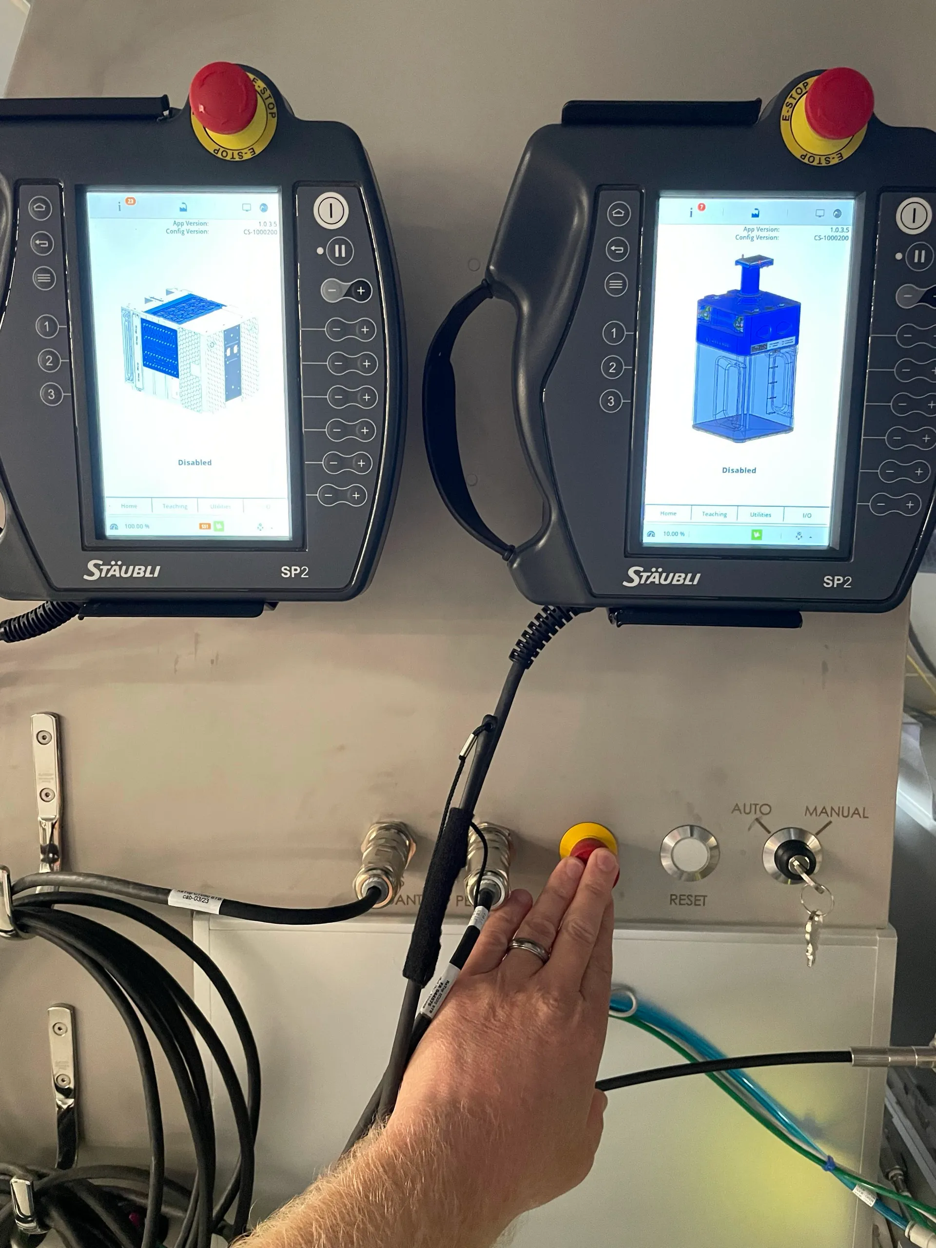 Two Stäubli robot teach pendants, each showing a 3D rendering of the equipment it controls, mounted next to a control panel with an auto/manual key switch and a hand pressing an E-stop button