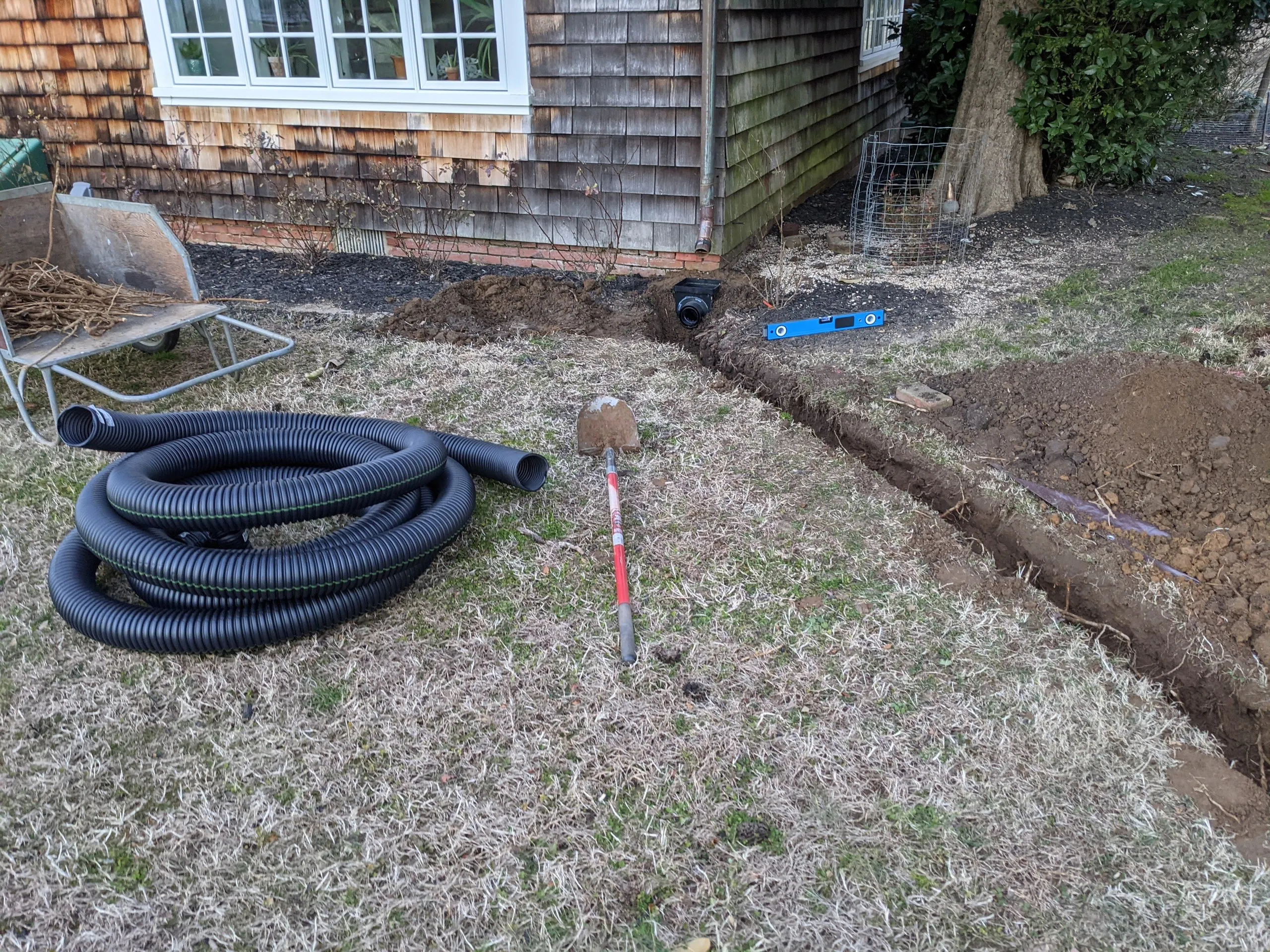 Coiled drain pipe and a shovel next to an open trench running from a catch basin along the foundation