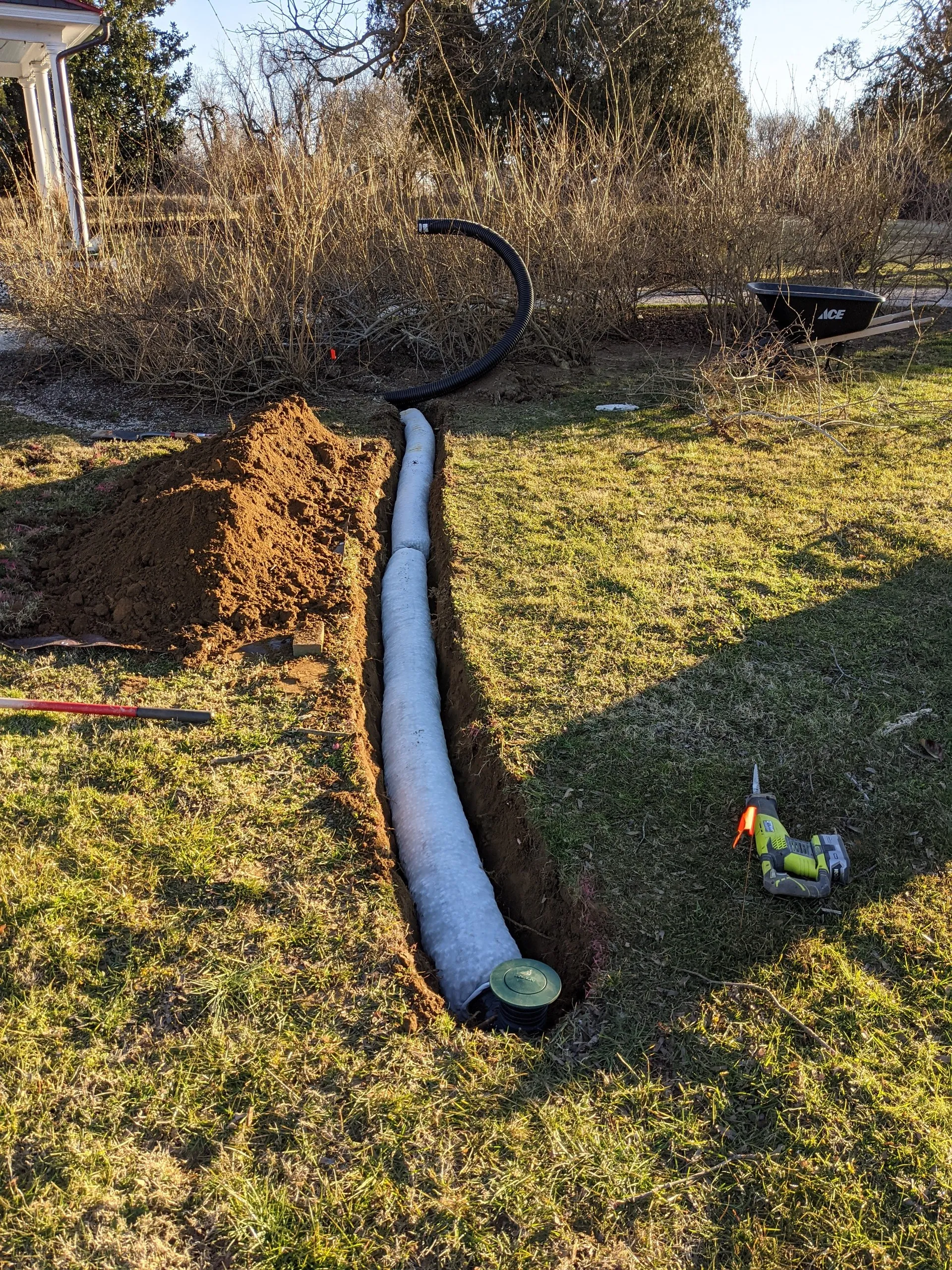 Long trench across the lawn with sock-wrapped pipe leading to a pop-up emitter, reciprocating saw left nearby